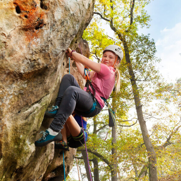 Young girl climbs a rock with harness in woodland at sunny day